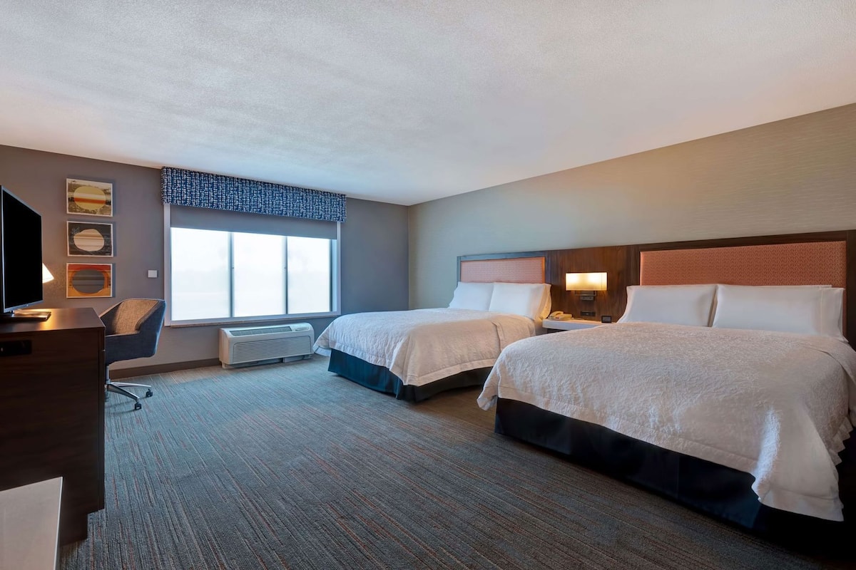 A hotel room with two beds, a desk, a TV, and a window with blinds.