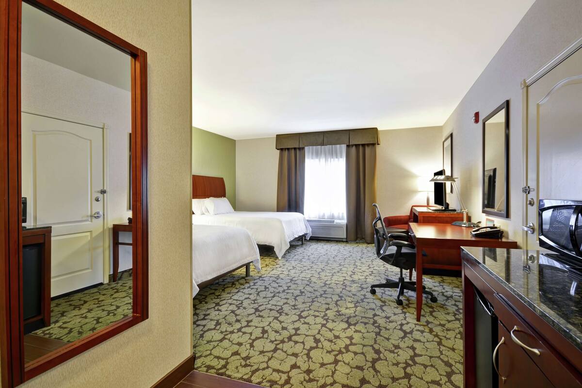 A hotel room with two beds, a desk with a chair, a microwave, and a large mirror.