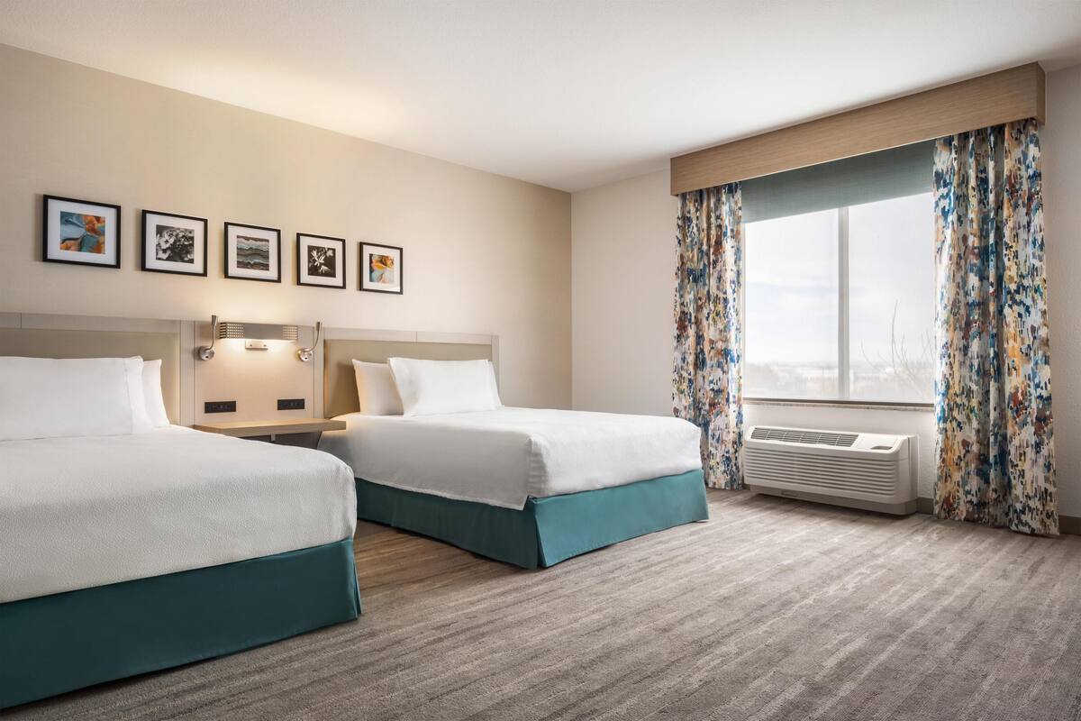 A hotel room with two beds, a large window with curtains, a wall-mounted air conditioner, and framed pictures on the wall.