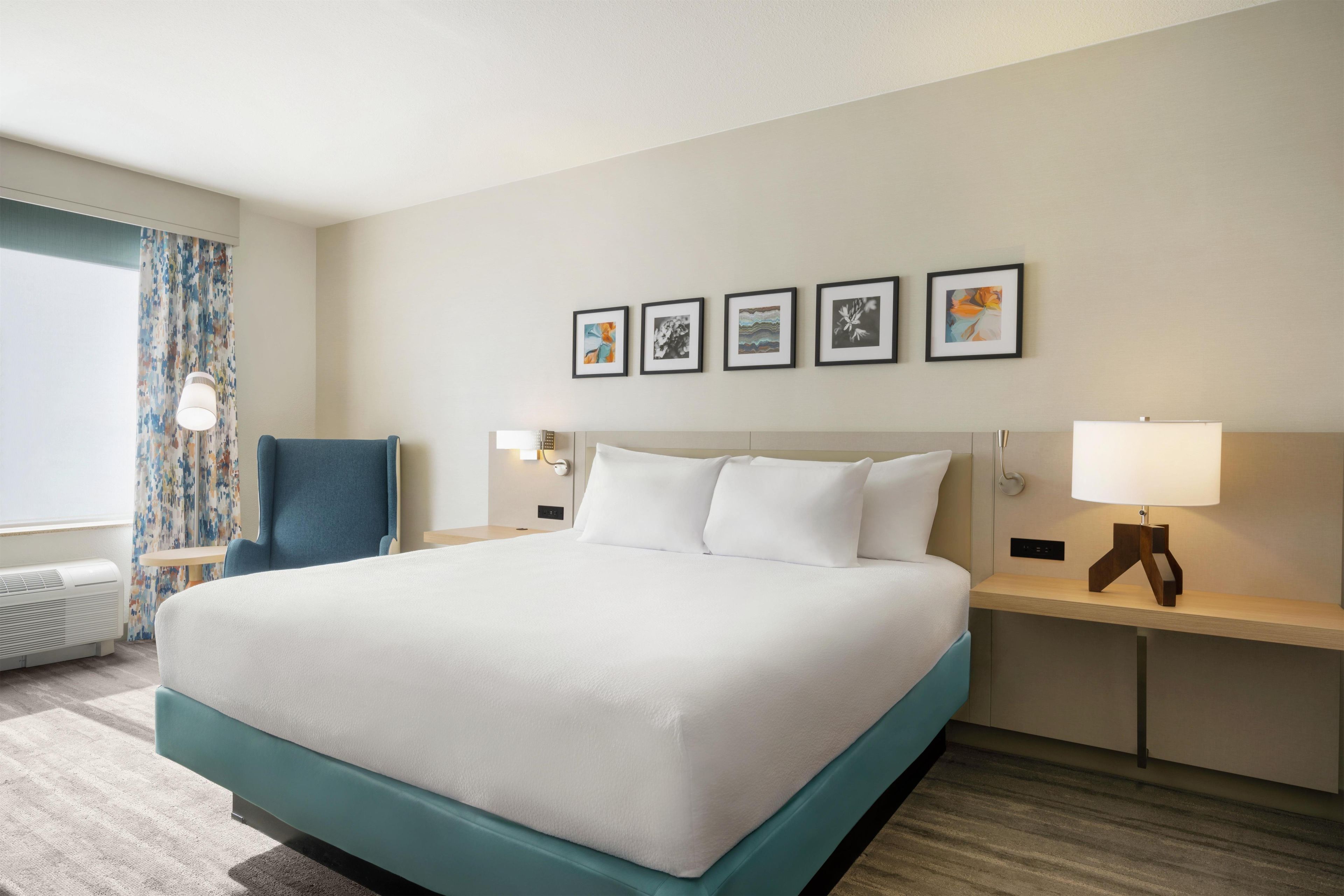 Quarto de hotel com uma cama grande, uma escrivaninha, uma cadeira e uma janela com cortinas.