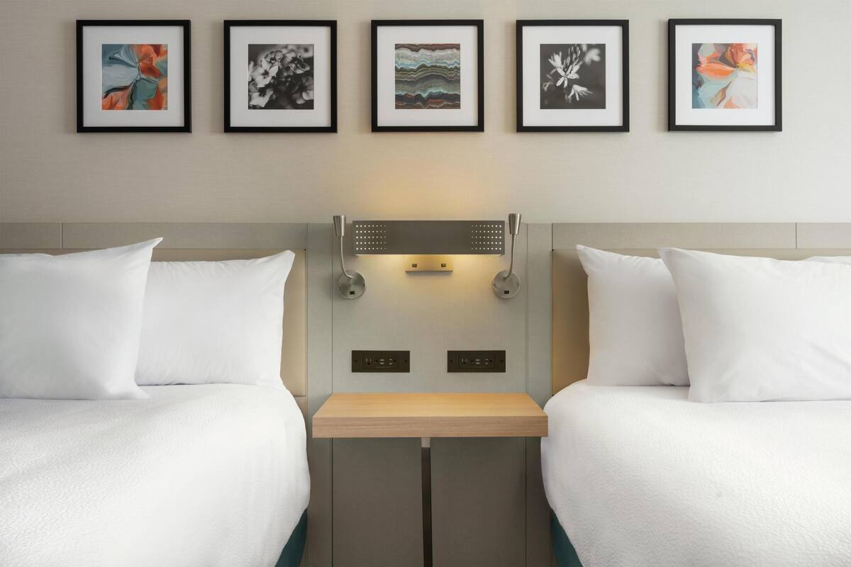 A hotel room with two beds, a bedside table, a wall-mounted light, and five framed artworks.