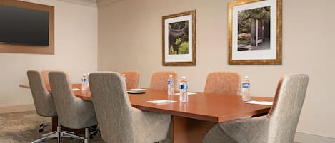 A conference room with a wooden table, six grey upholstered chairs, two framed pictures on the wall, and a flat-screen TV on the left.