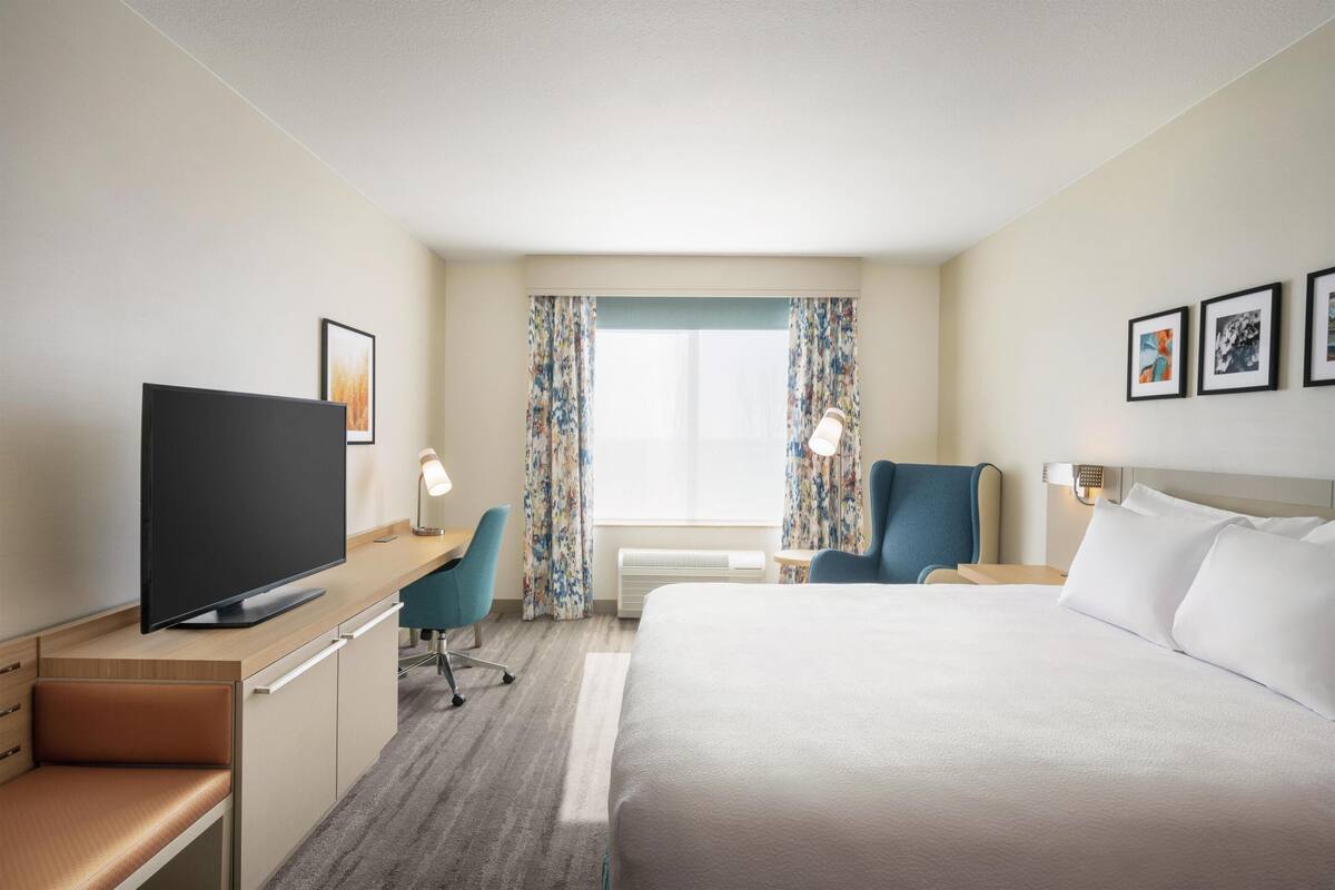 A hotel room with a bed, desk, TV, chair, and window with curtains.