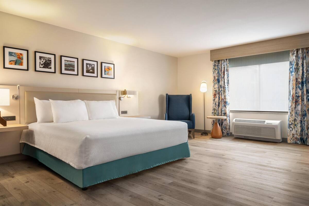 A hotel room with a bed, a chair, a lamp, a window with curtains, and a wall with framed pictures.
