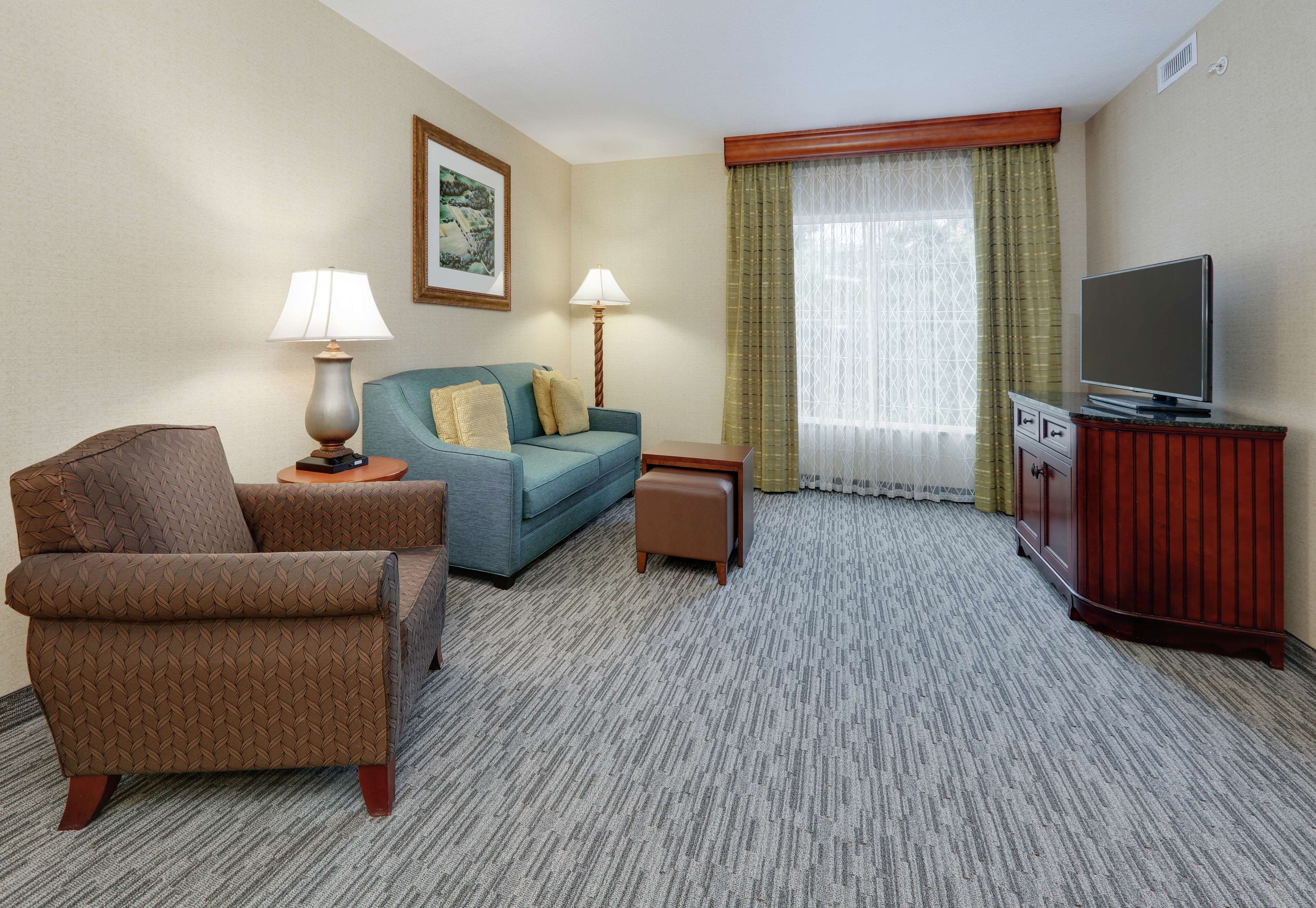 Una habitación de hotel con un sillón marrón, un sofá azul, una mesita con una lámpara, un televisor sobre un mueble de madera y un ventanal con cortinas.