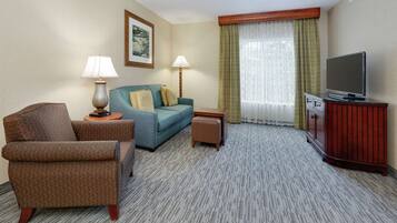 Una habitación de hotel con un sillón marrón, un sofá azul, una mesita con una lámpara, un televisor sobre un mueble de madera y un ventanal con cortinas.