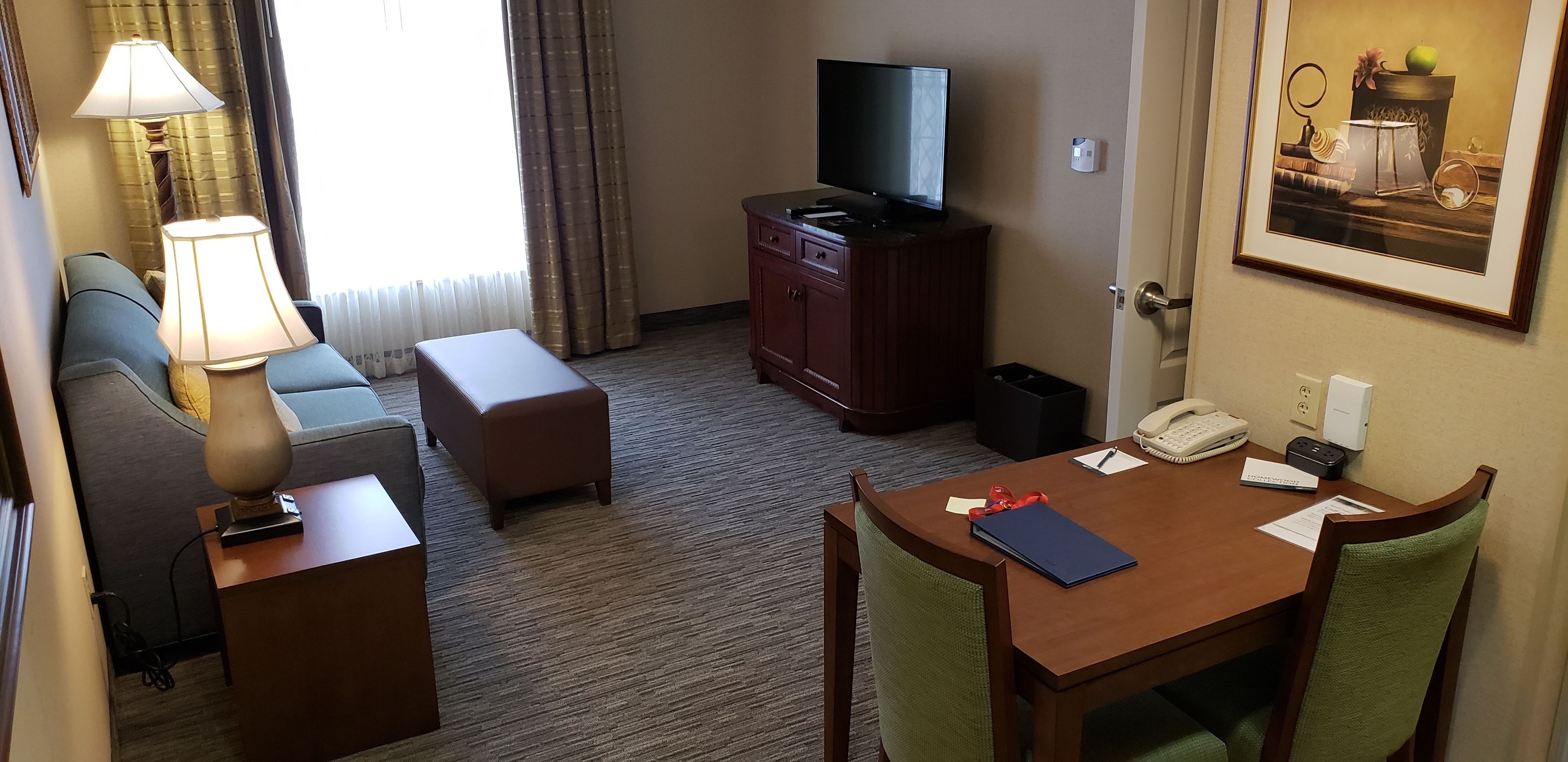 Een hotelkamer met een bank, een klein tafeltje, een televisie op een kast, een schilderij aan de muur en een raam met gordijnen.