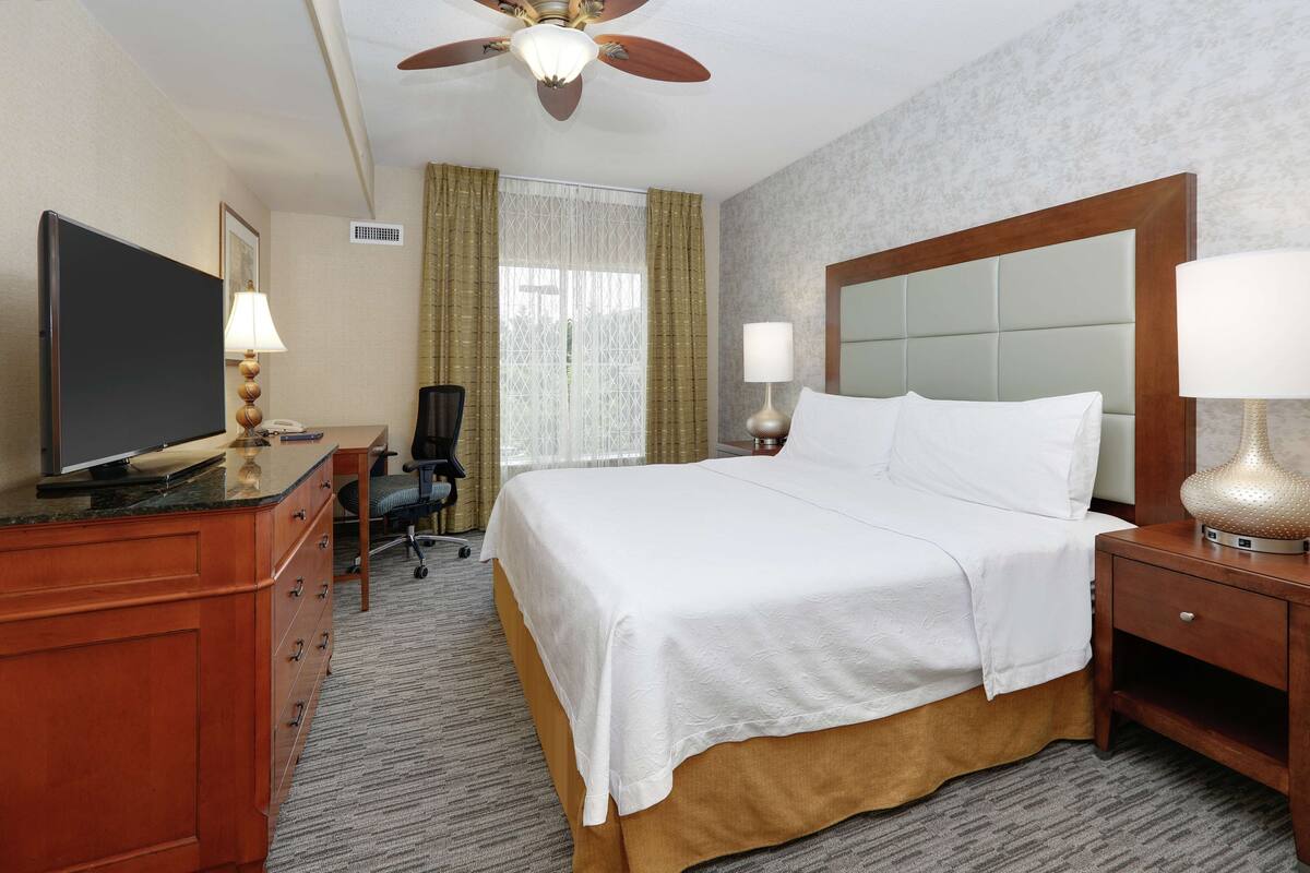 Una habitación de hotel con cama, escritorio, televisor y ventilador de techo.