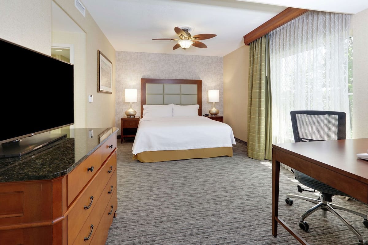 Una habitación de hotel con cama, escritorio, televisión, ventilador de techo y ventana con cortinas.