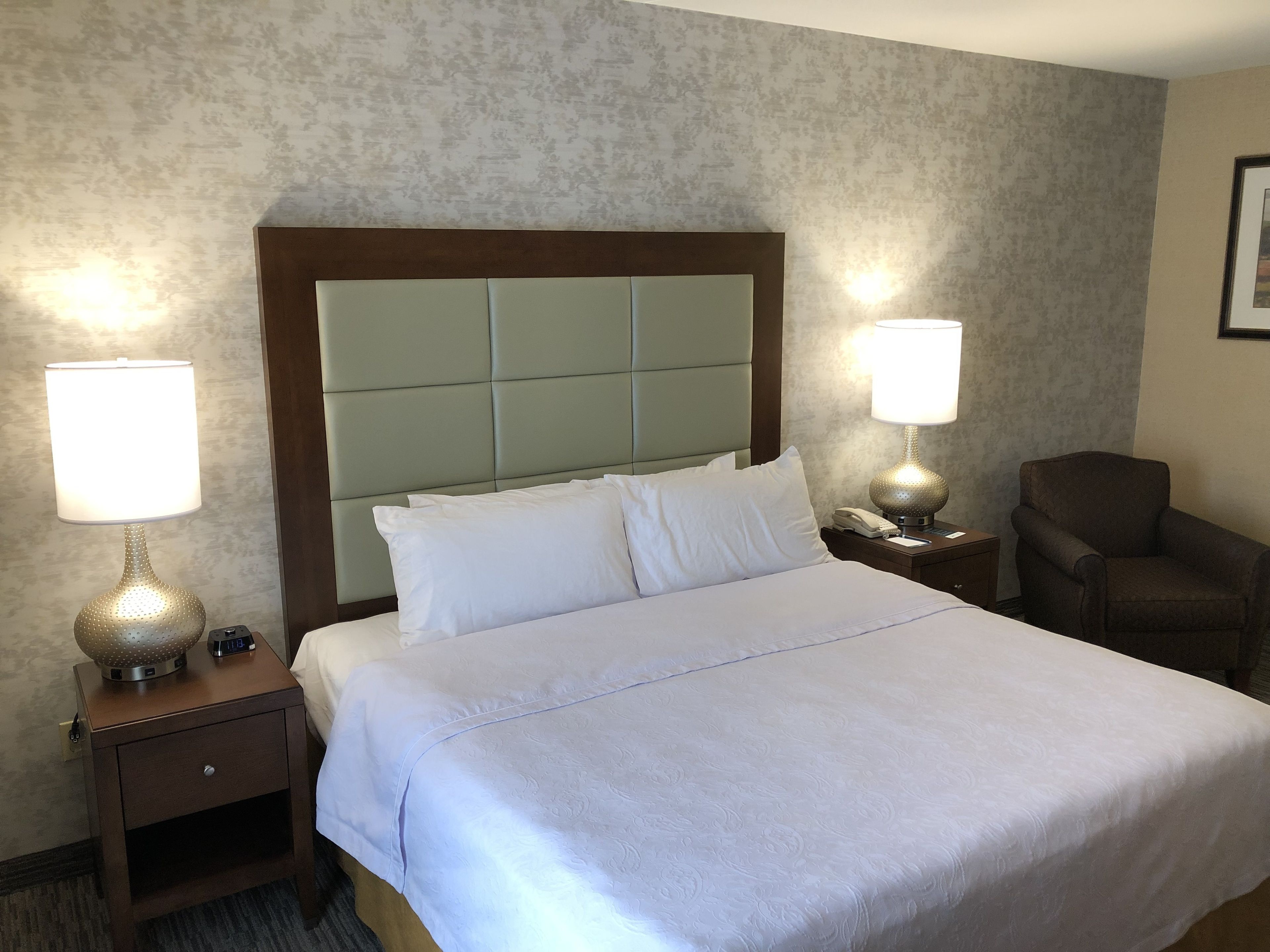A hotel room with a large bed, two bedside lamps, a chair, and a nightstand with a phone and a clock.