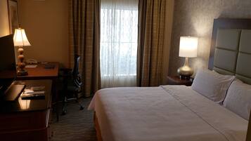 A hotel room with a bed, a desk with a lamp, a chair, and a window with curtains.