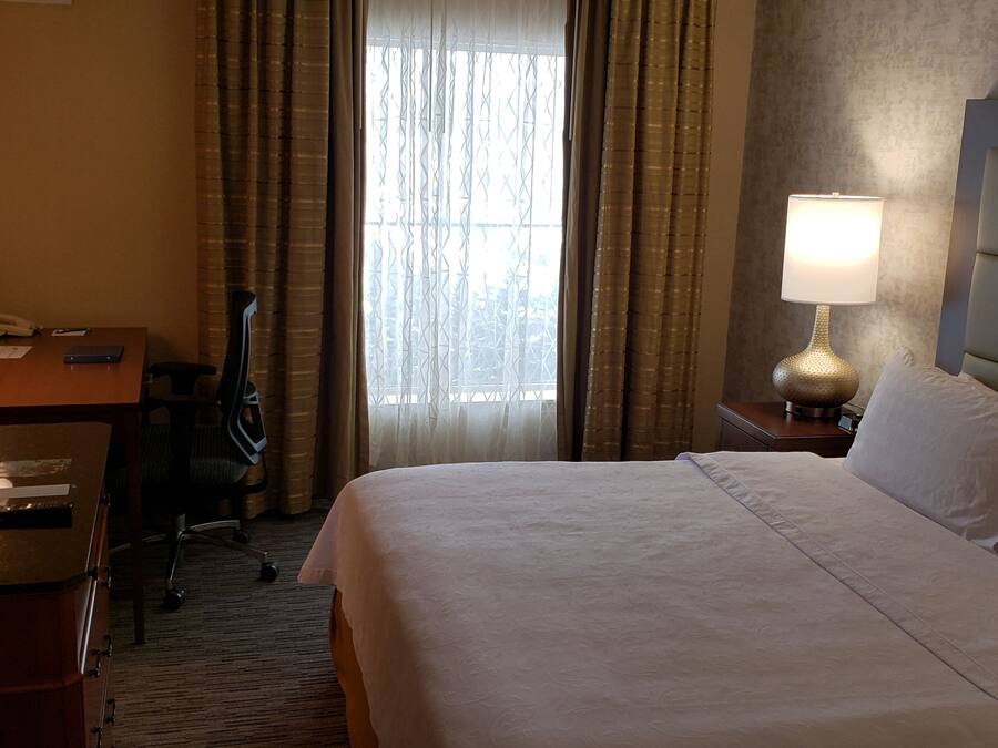 A hotel room with a bed, a desk with a lamp, a chair, and a window with curtains.