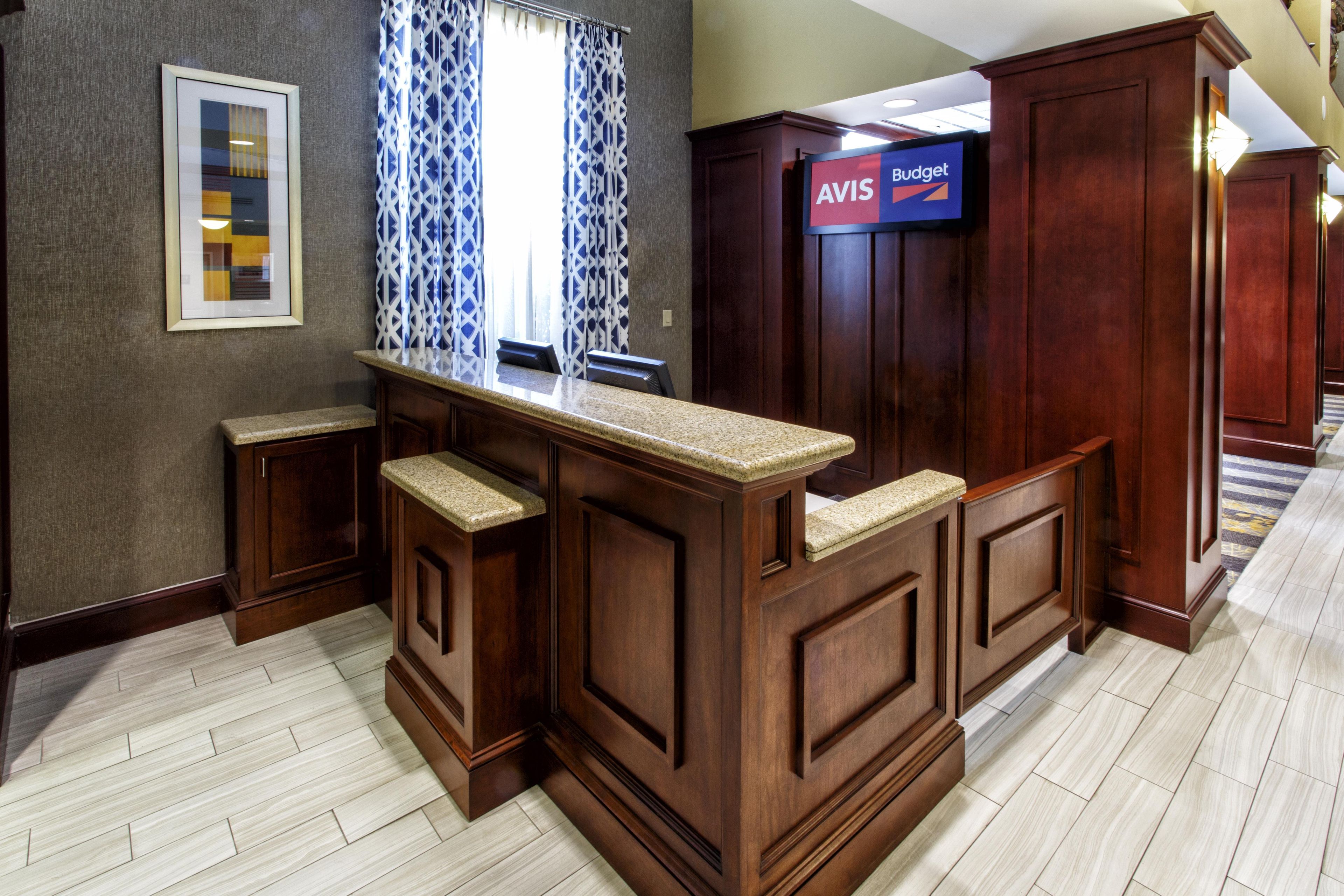 A hotel lobby with a wooden reception desk, a framed artwork on the wall, and a television screen displaying the AVIS Budget logo.