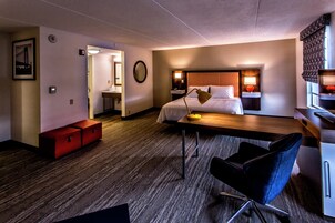 A hotel room with a bed, bedside table, lamp, chair, desk, and a small red cabinet.