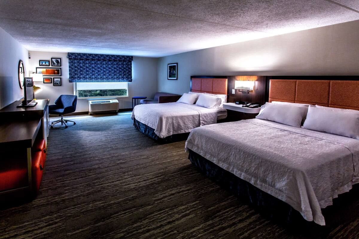 A hotel room with two beds, a desk, a chair, a lamp, and a window with a view.