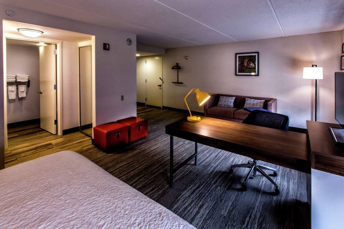 A hotel room with a bed, a desk, a chair, a sofa, a red bench, and a lamp.