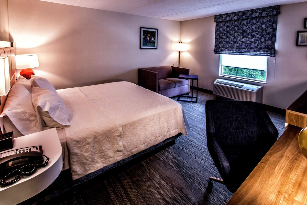 A hotel room with a bed, a desk, a chair, a lamp, and an air conditioning unit.
