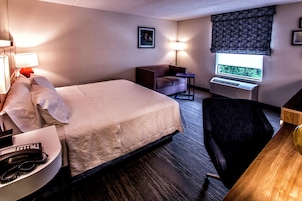 Habitación de hotel con cama, escritorio, silla, lámpara, un cuadro en la pared y aire acondicionado.