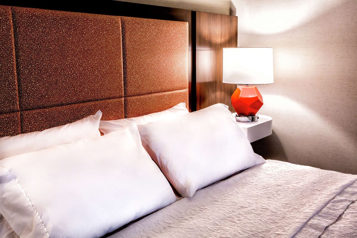 A neatly made bed with white linens and pillows, a wooden headboard, and a bedside table with a red lamp.