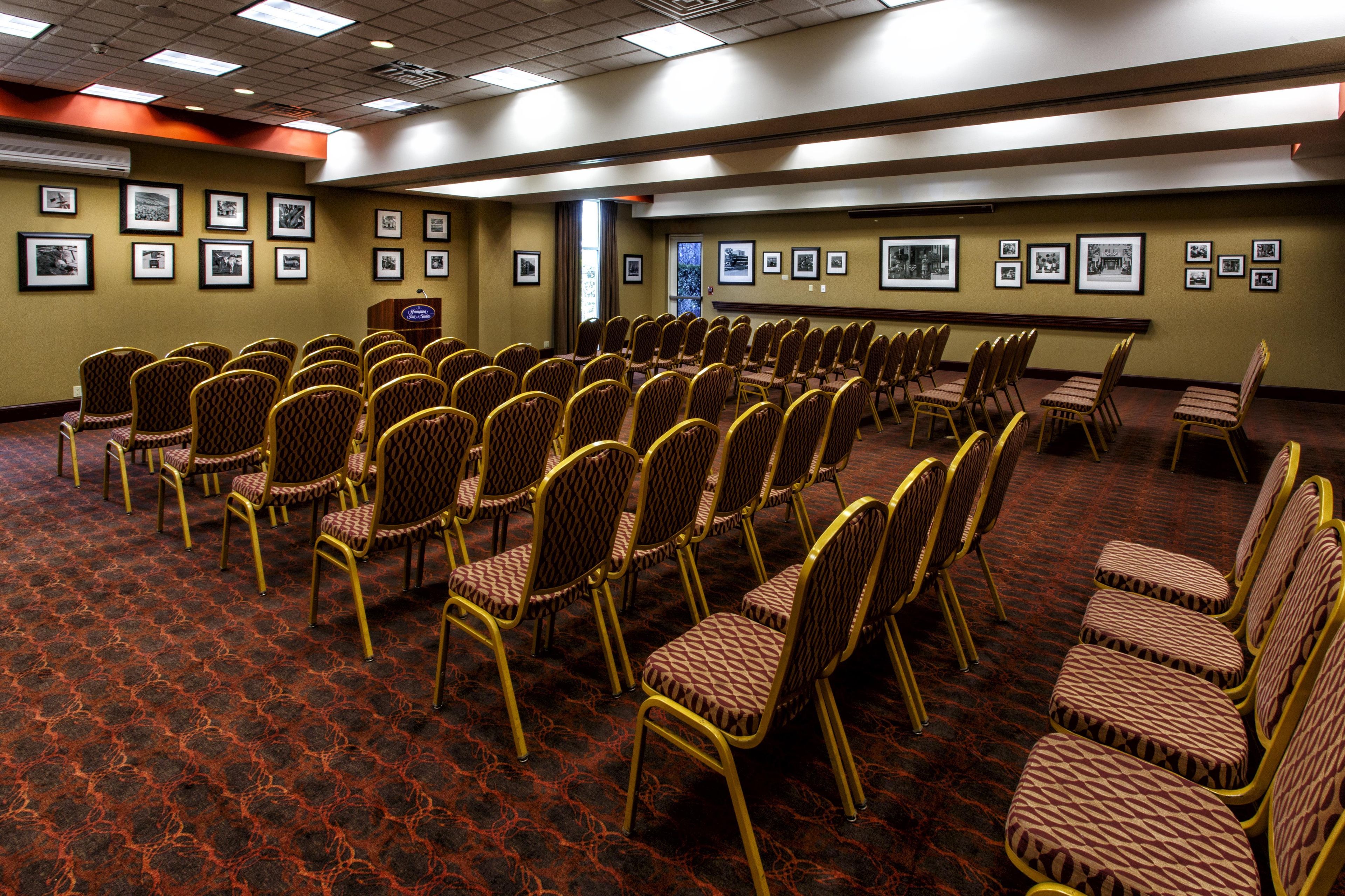 A conference room with rows of chairs and a podium, walls adorned with framed pictures, and a carpeted floor.