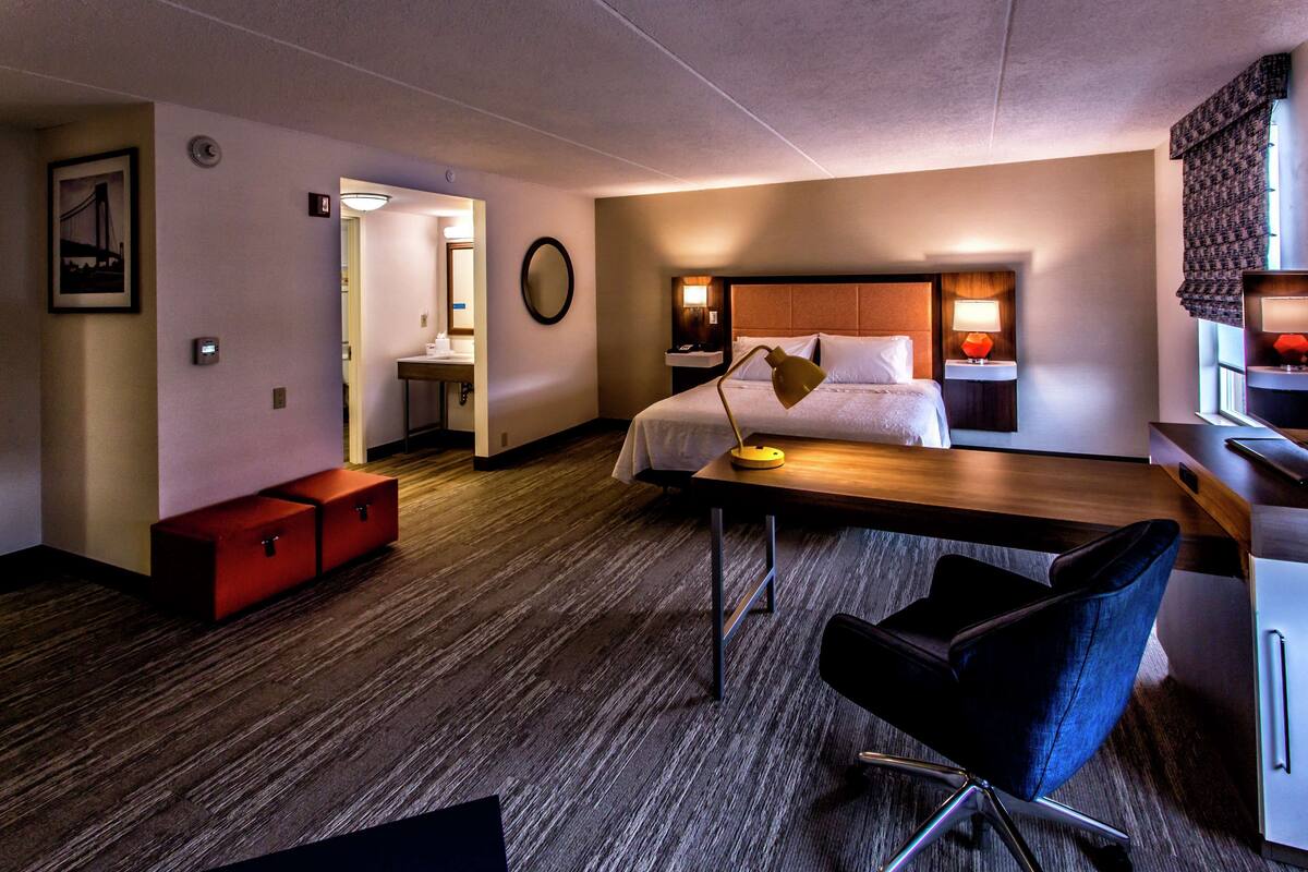 A hotel room with a bed, bedside tables, a desk, a chair, a wardrobe, and a bathroom visible through an open door.