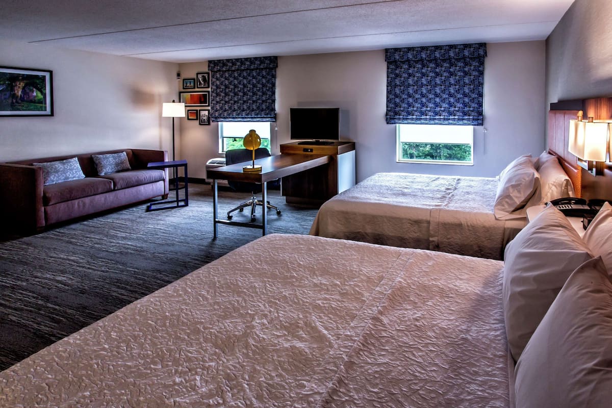 A hotel room with two beds, a desk, a sofa, a TV, and a window with a view.