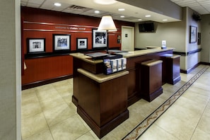 A reception desk with framed pictures on the wall, a counter with brochures, and a tiled floor. - Hampton Inn & Suites Staten Island (Staten Island)