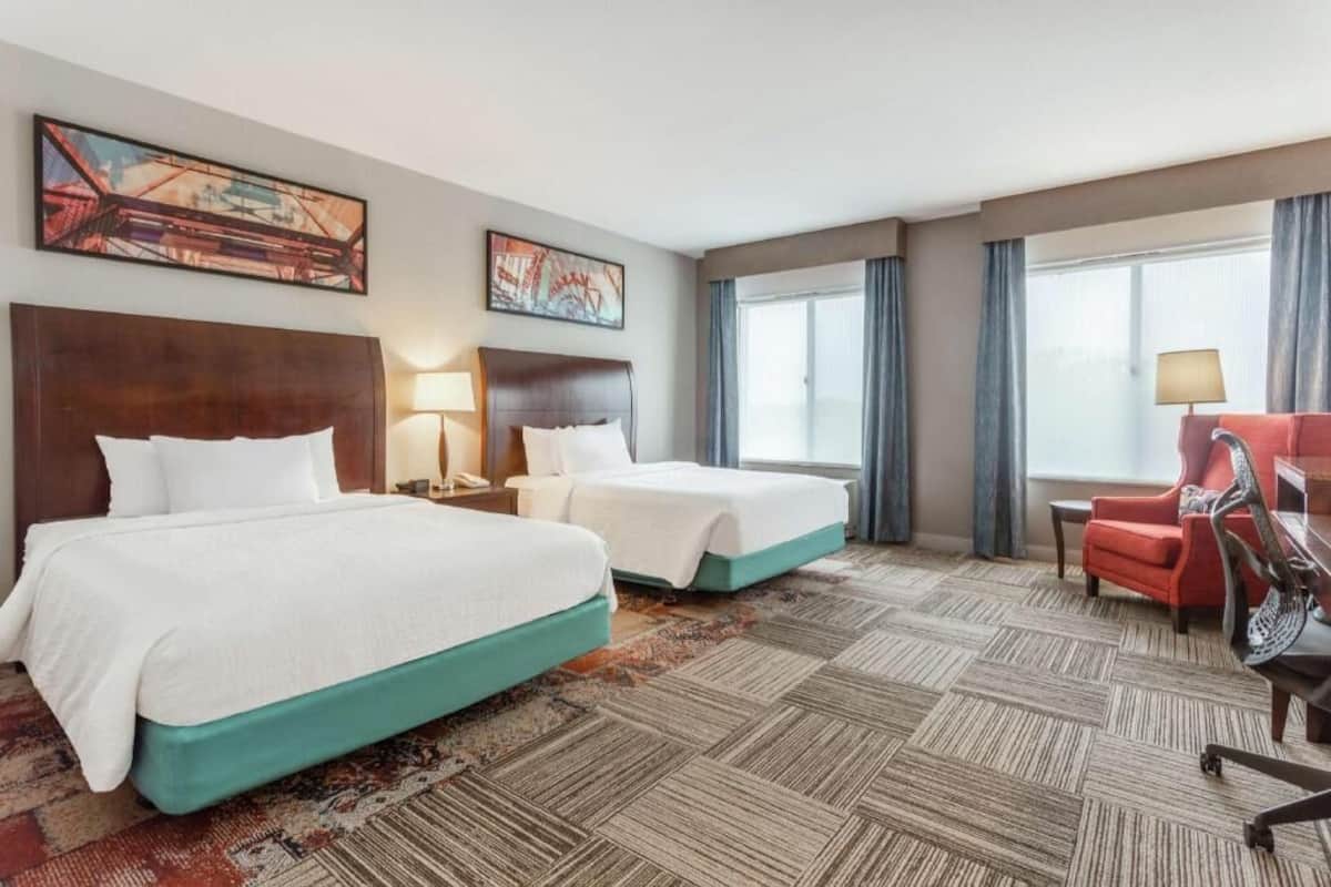 A hotel room with two beds, a red chair, a desk, and a large window with curtains.