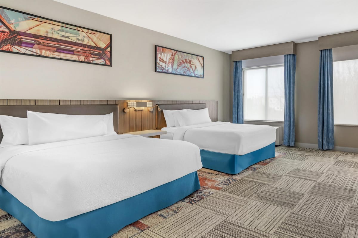 A hotel room with two beds, a large window with curtains, and artwork on the walls.