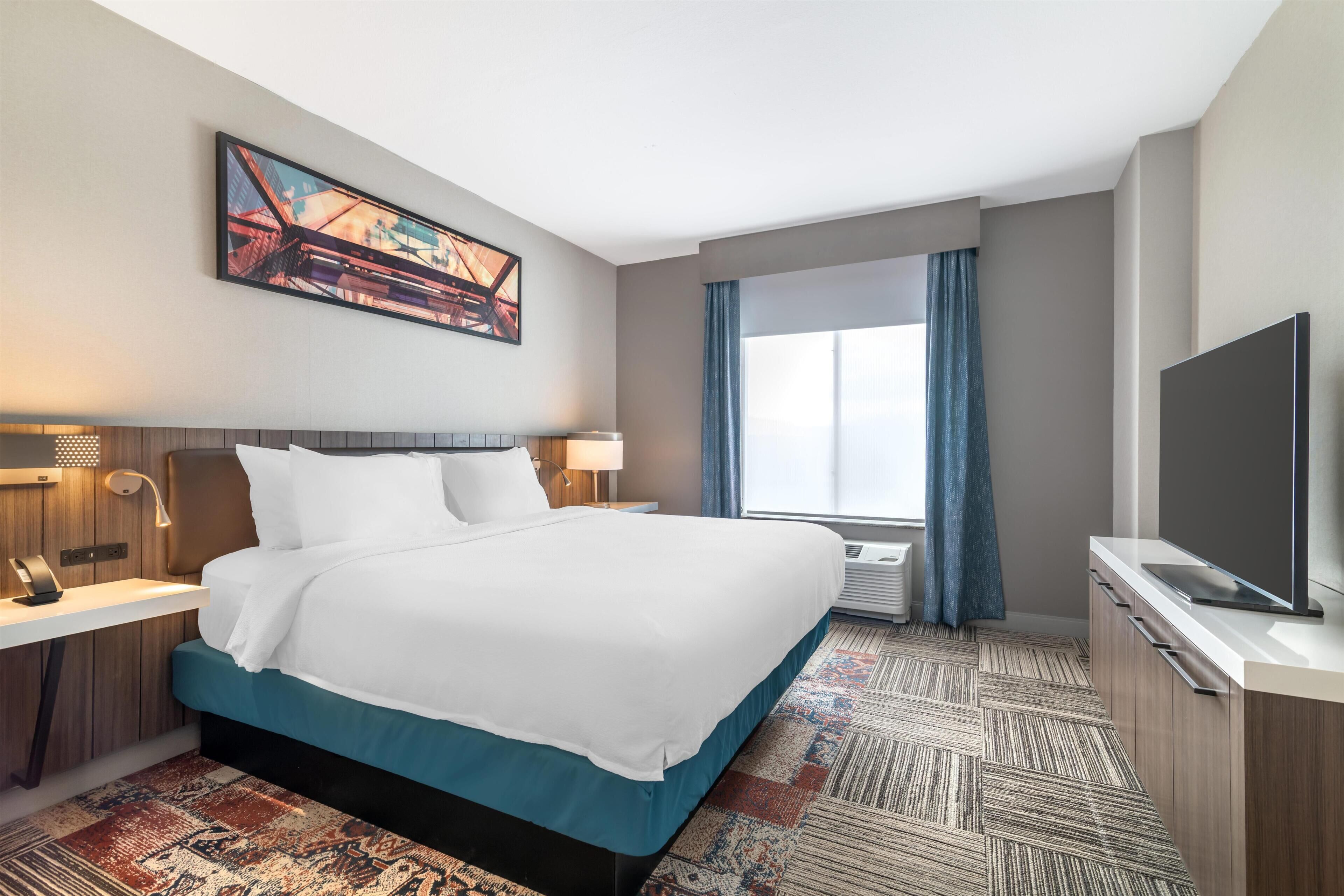 A hotel room with a bed, bedside tables, a TV, a large window with curtains, and a painting on the wall.