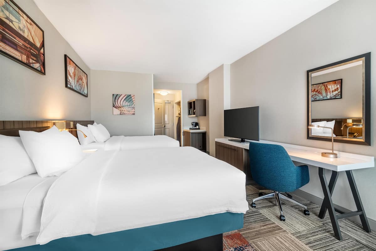 A hotel room with two beds, a desk, a chair, a TV, and artwork on the walls.