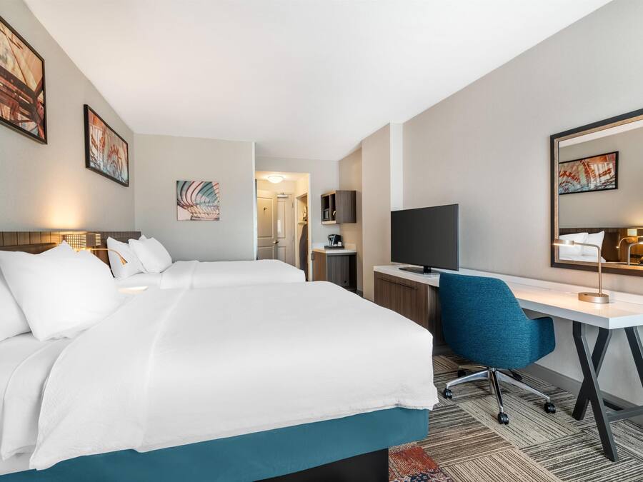 A hotel room with two beds, a desk, a chair, a TV, and artwork on the walls.