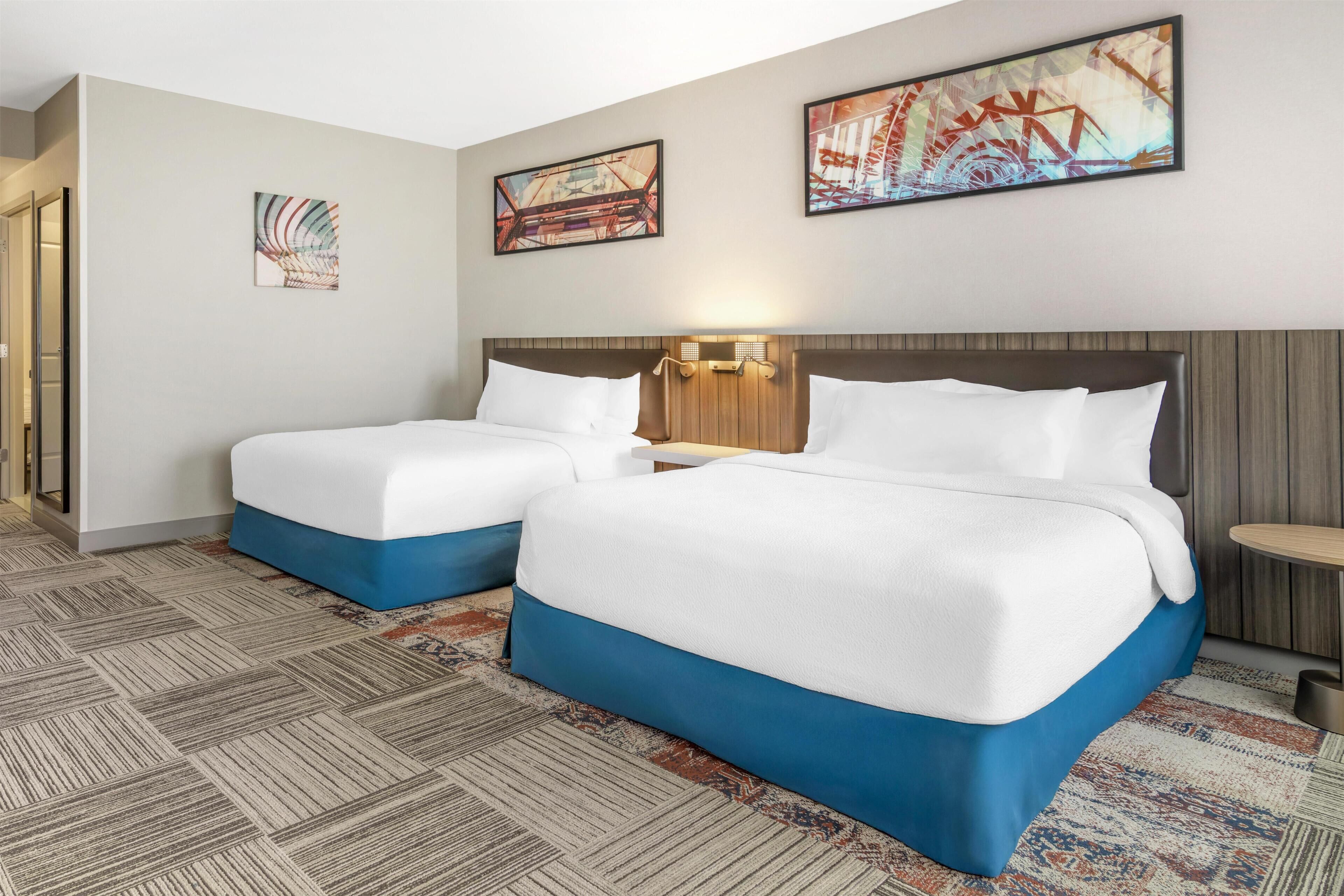 A hotel room with two beds, a wooden headboard, a nightstand, and a wall-mounted lamp.