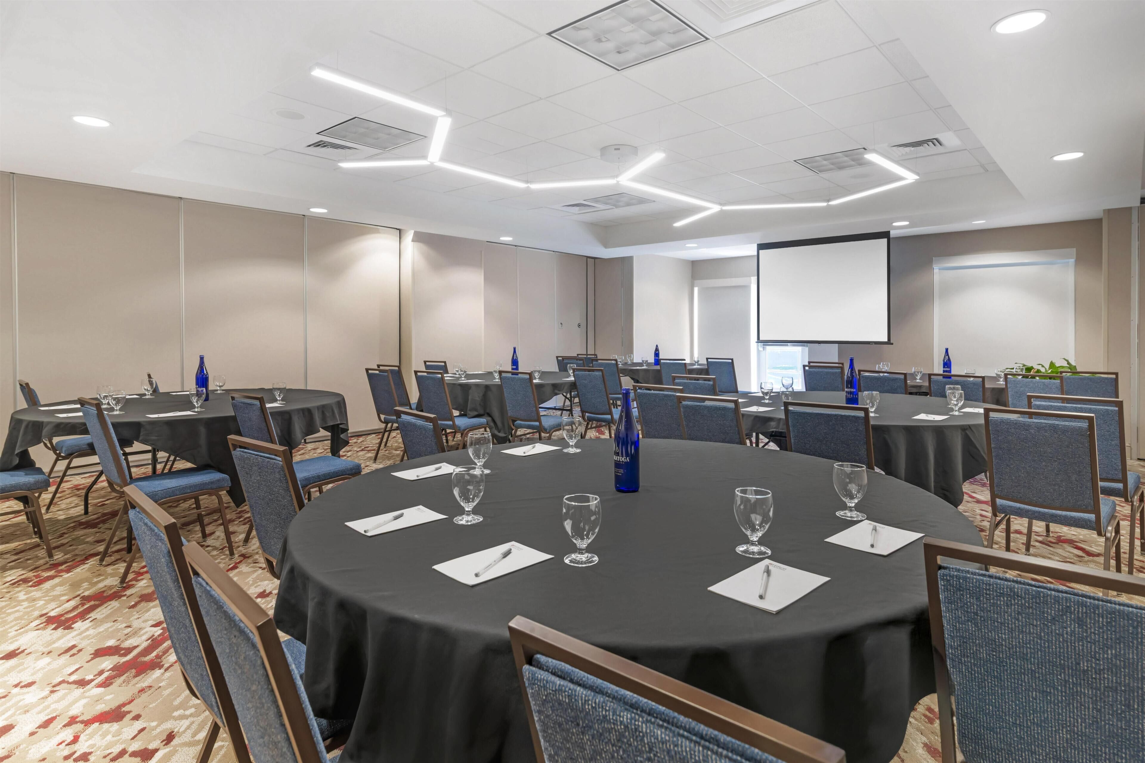 Un salón de conferencias con mesas redondas, sillas y una pantalla de proyector.
