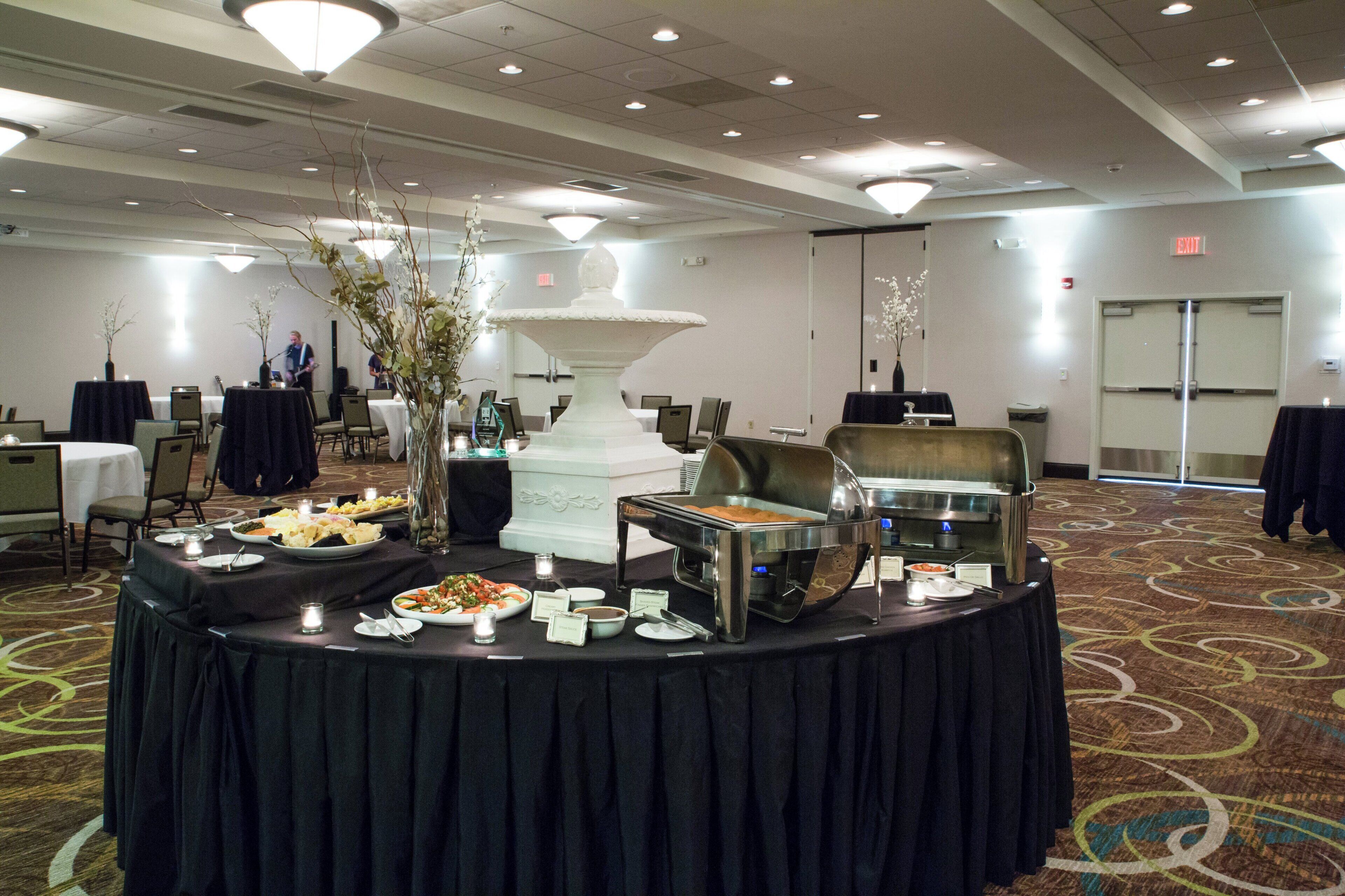 A banquet hall with round tables set for a meal, a buffet area with various food options, and a stage for presentations.