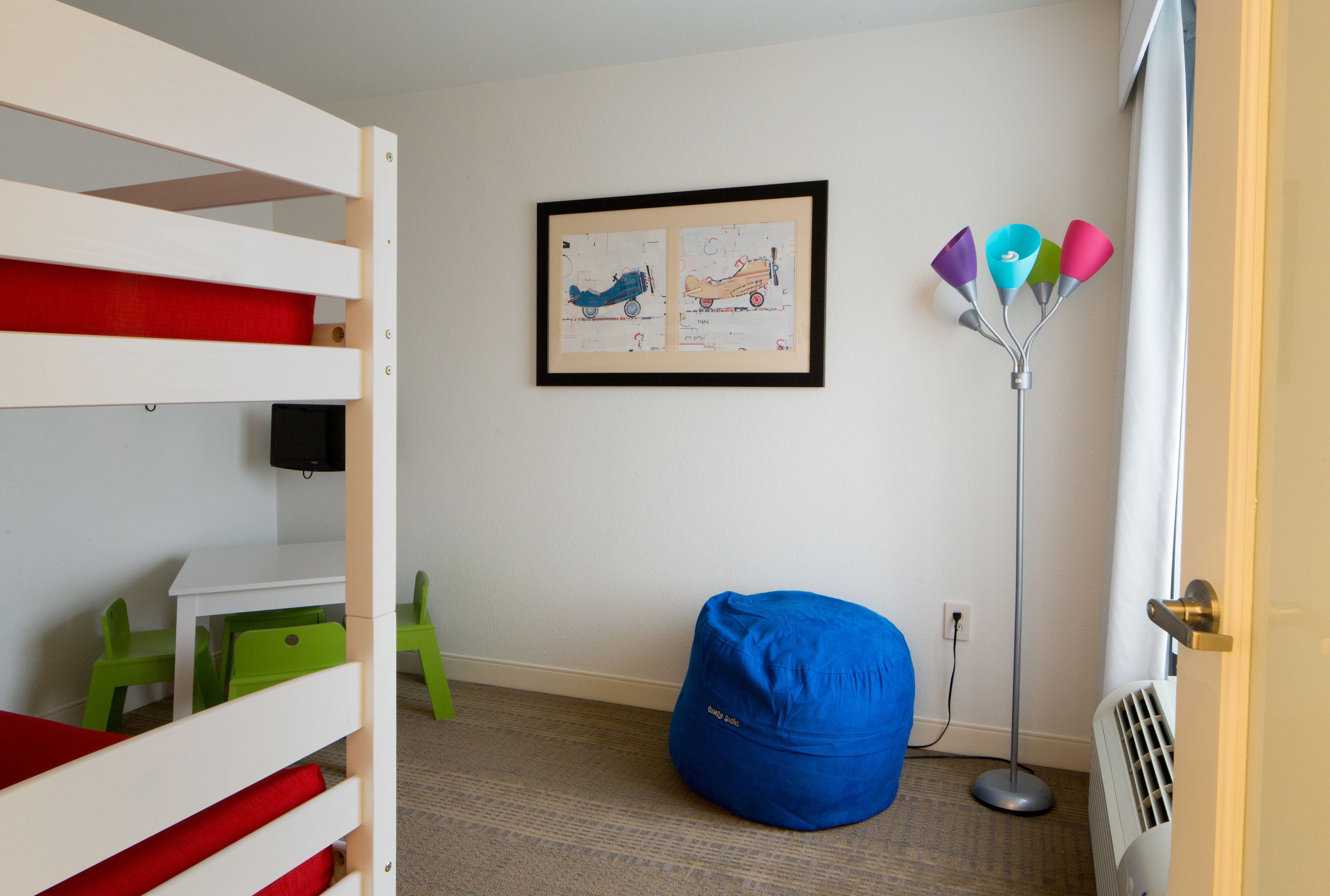 Habitación infantil con literas, un sillón de peluche, un cuadro enmarcado y una lámpara de pie.