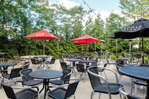 Outdoor seating with tables and chairs, red umbrellas, and greenery in the background. - Holiday Inn Bloomington-Airport by IHG (Bloomington)