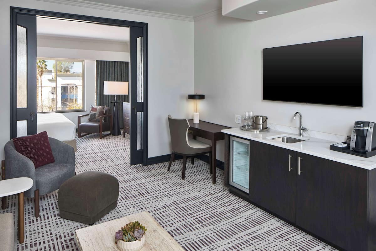 A modern hotel room with a kitchenette, a flat-screen TV, a dining area with a chair and table, and a view through an open door.