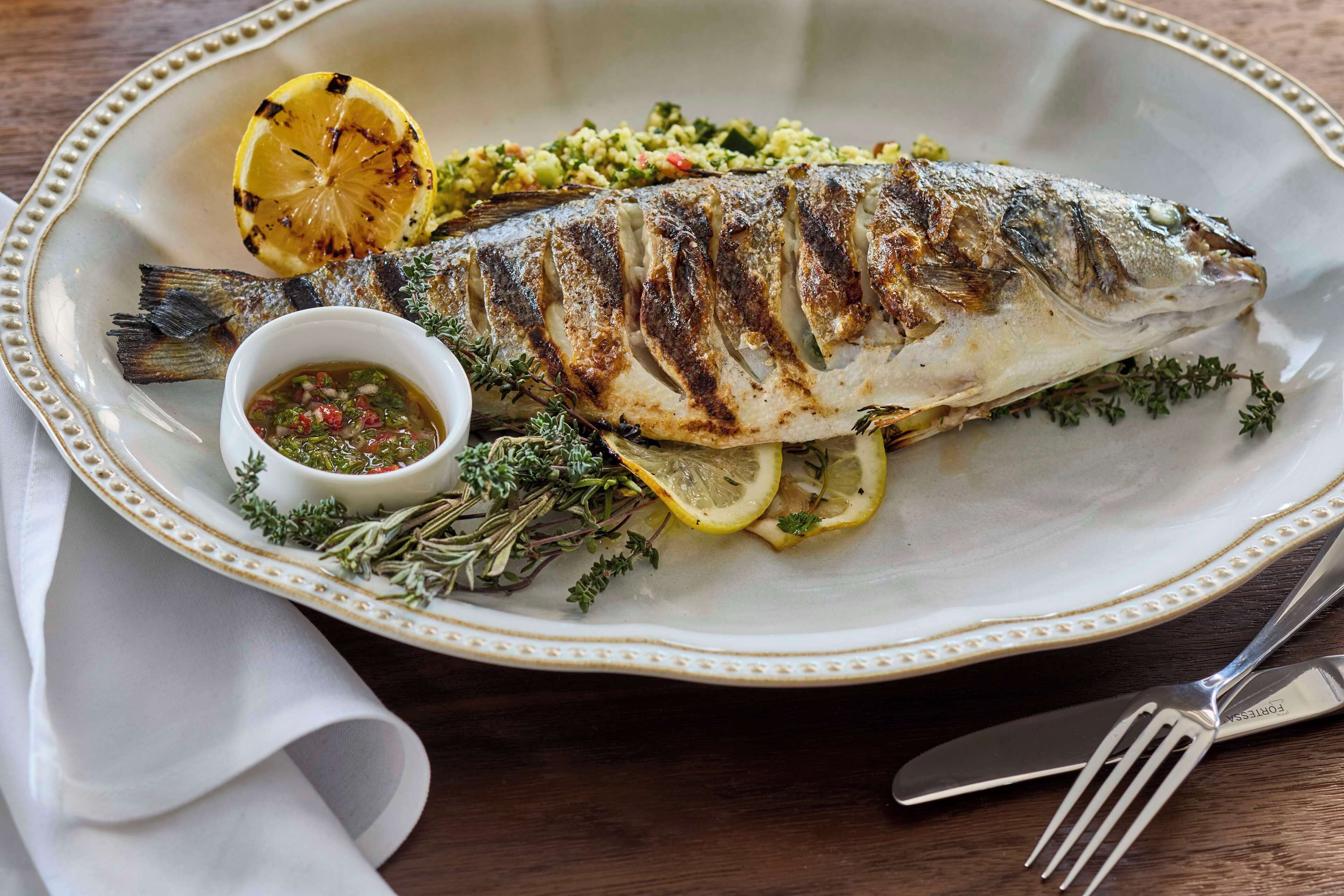 A plate with grilled fish, lemon slices, herbs, and a small bowl of sauce.
