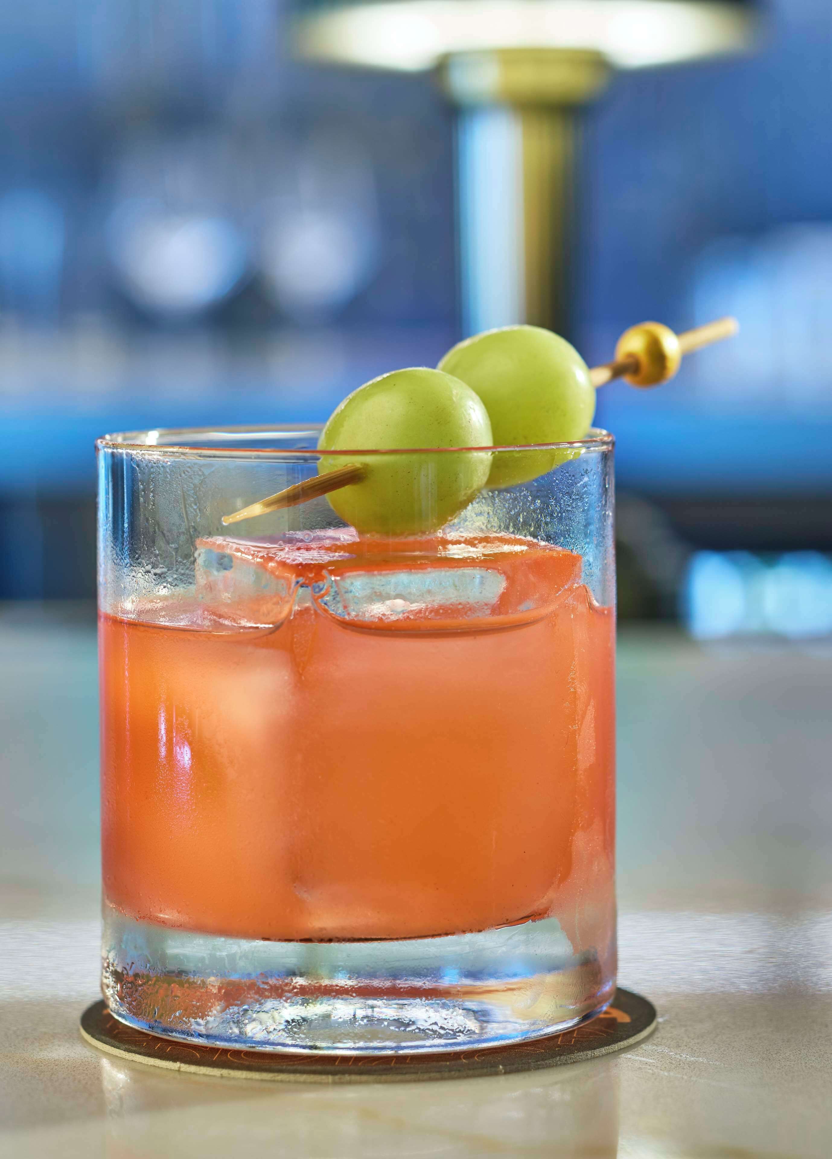 A cocktail glass with a clear liquid, ice cubes, and a green fruit garnish on a coaster.