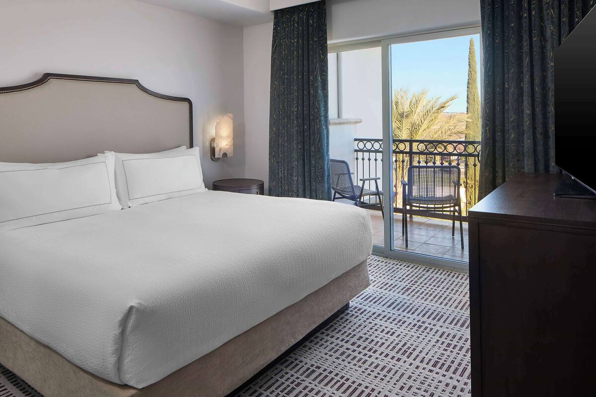 A hotel room with a large bed, a small table, a chair, a TV, and a balcony with a view of palm trees.