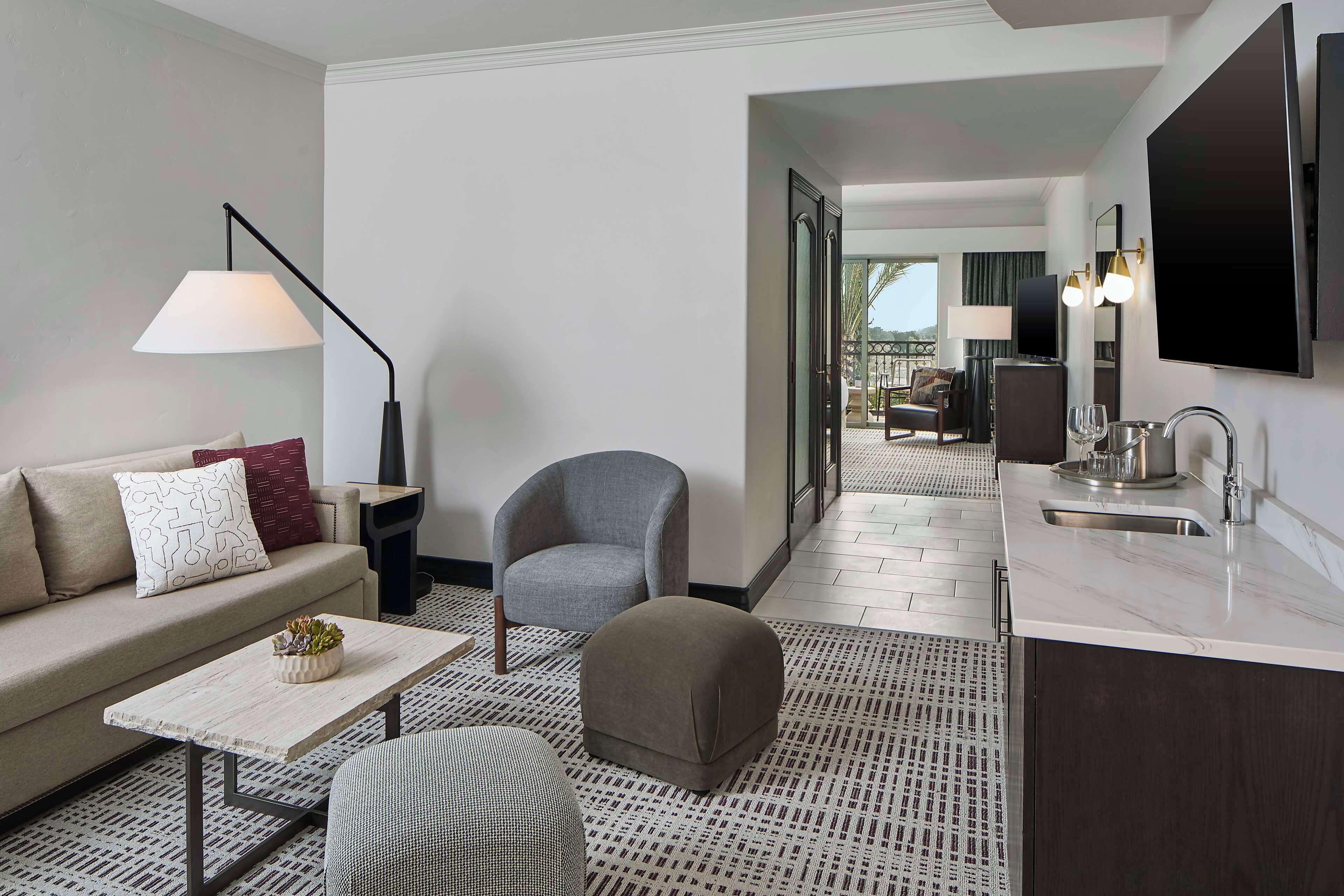 Una habitación de hotel moderna con sofá, sillón, reposapiés, mesa de centro y una zona de cocina con lavamanos y encimera.