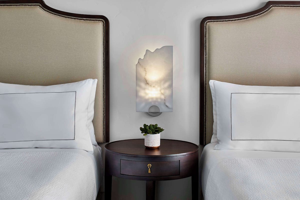 A hotel room with two beds, a nightstand with a plant, and a wall-mounted light fixture.