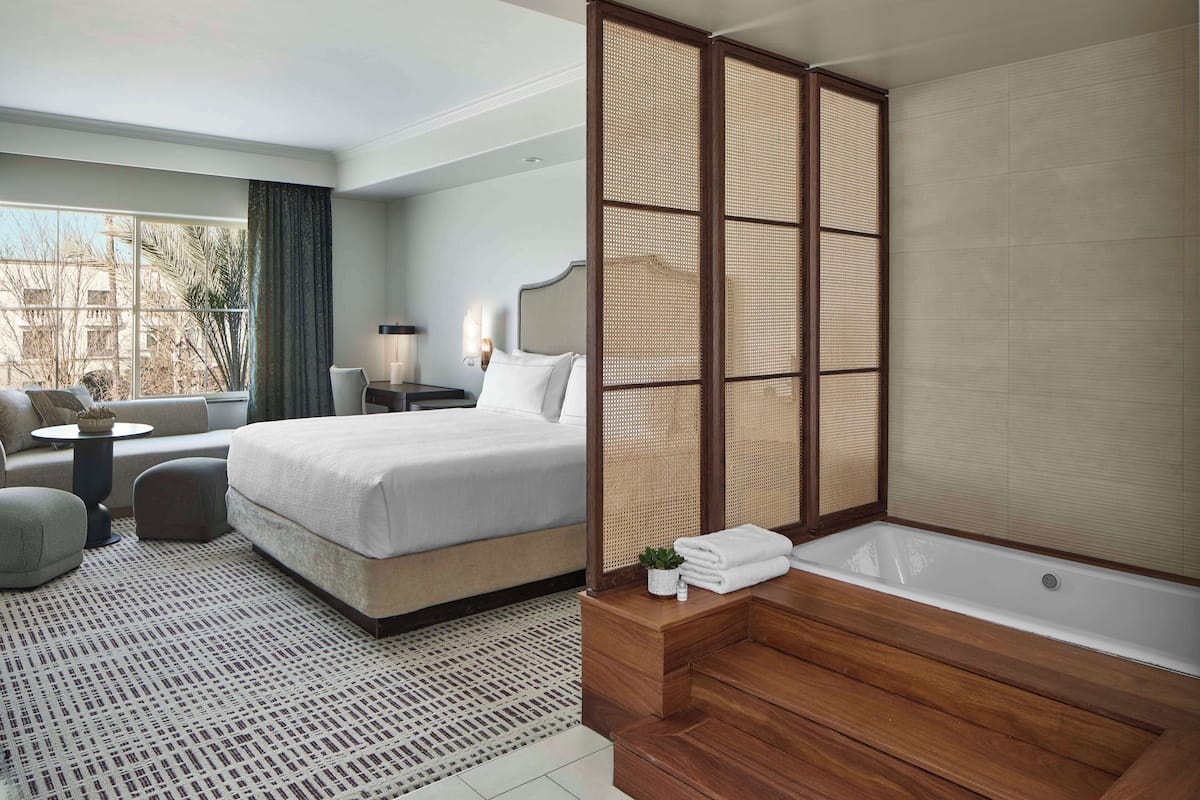 A modern hotel room with a large bed, a sitting area with a sofa and armchair, a small table, a large window with a view, and a separate bathroom with a bathtub and wooden vanity.