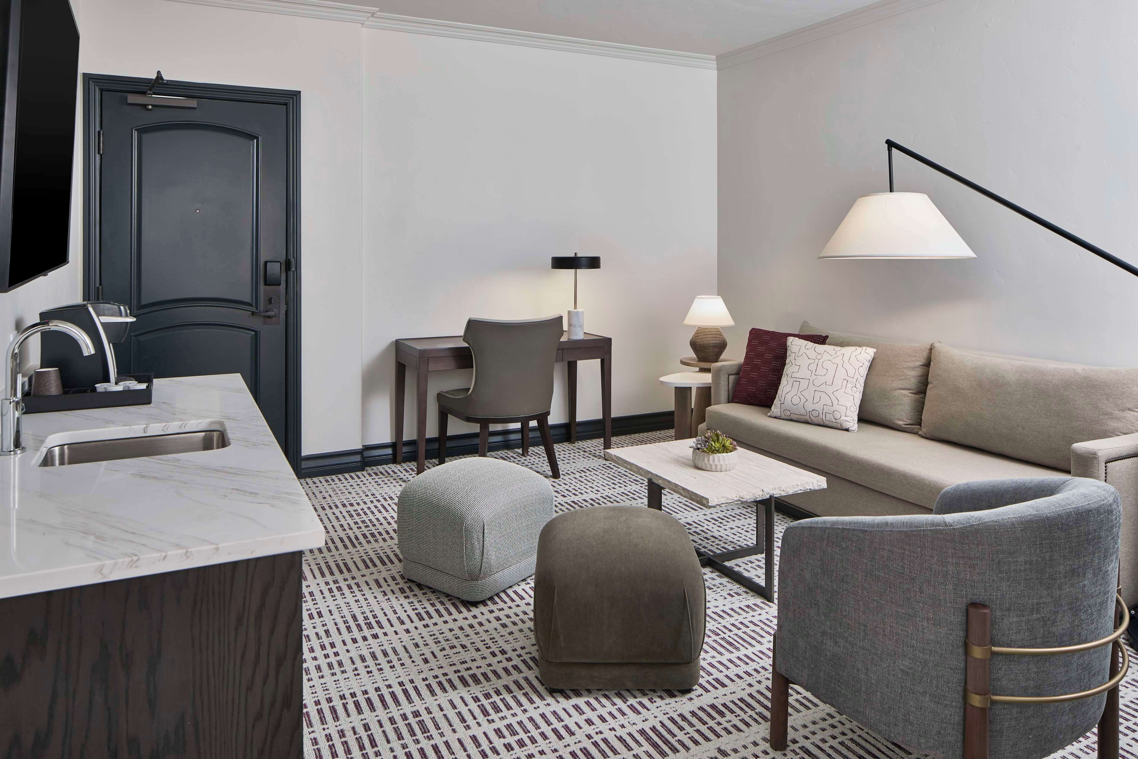 A modern hotel room with a marble kitchen island, a grey sofa with a patterned rug, and a minimalist decor style.