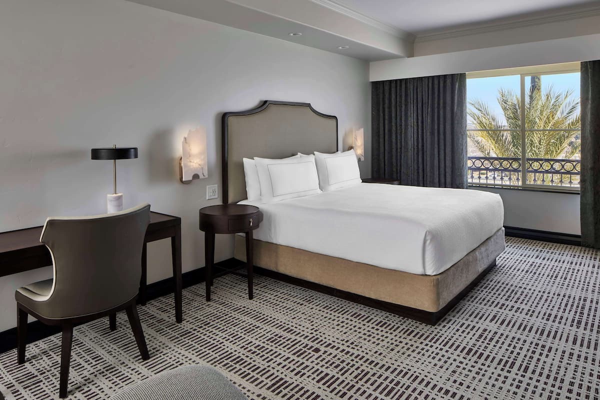 A hotel room with a large bed, a desk, a chair, and a view of palm trees through the window.