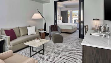 Hotellihuone, jossa on sohva, pieni pöytä, tuoli, sänky ja pieni keittiönurkkaus.