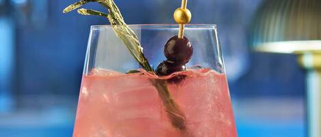 Um cocktail rosa num copo com haste, decorado, apoiado num balcão de bar.