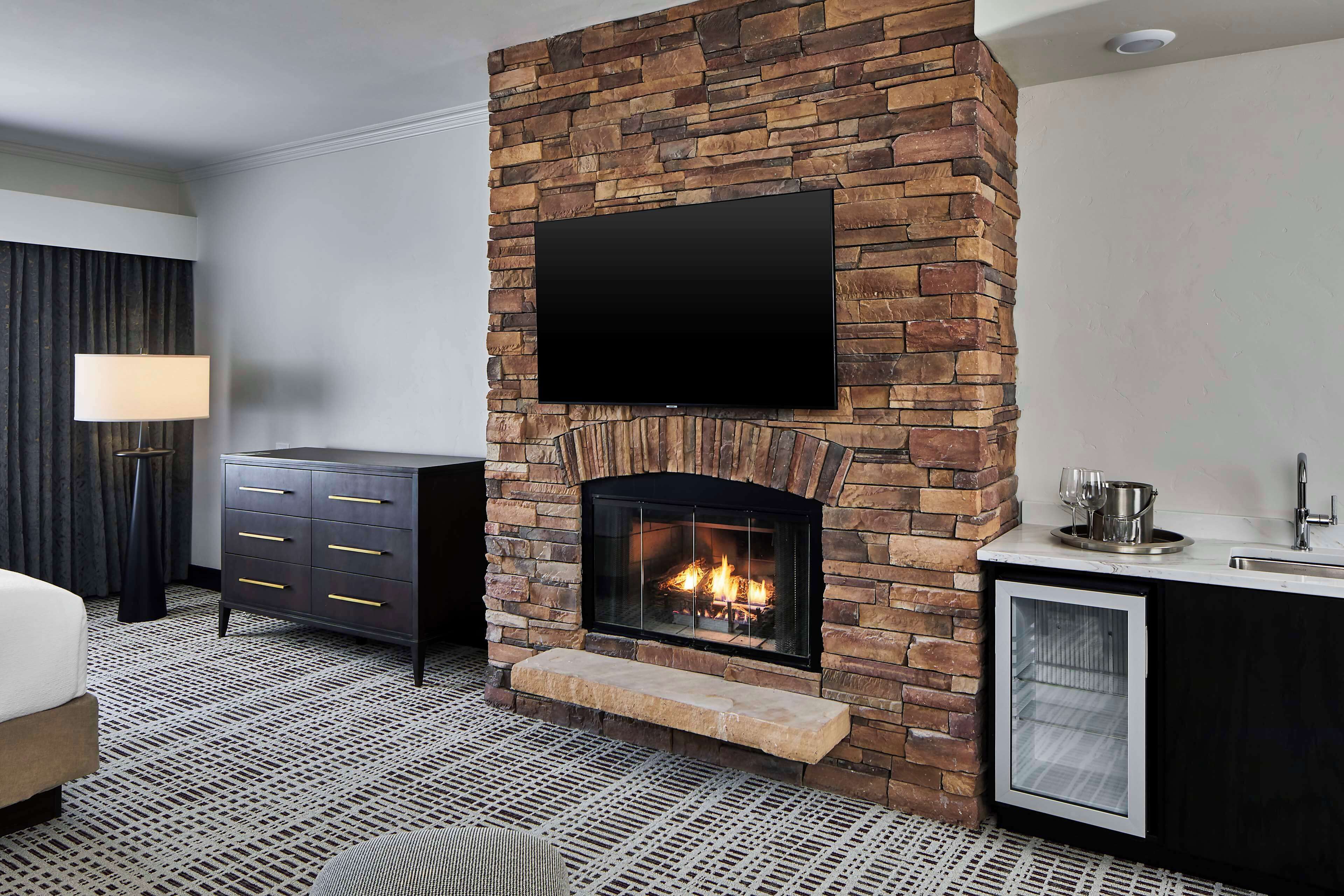 A modern hotel room with a stone fireplace, a flat-screen TV mounted above it, a dark-colored dresser, and a small kitchenette area with a sink and a refrigerator.