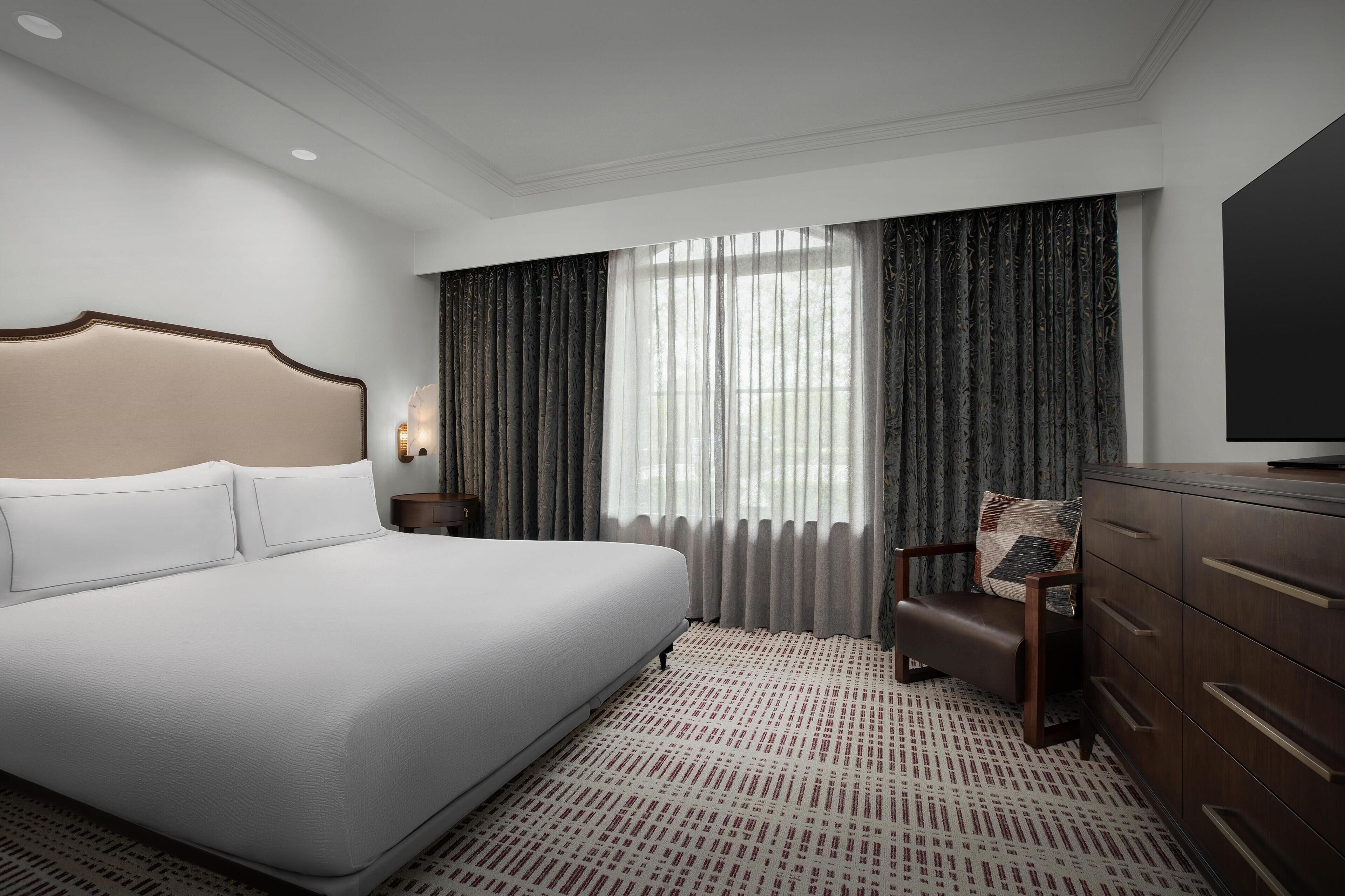 Une chambre d’hôtel avec un grand lit, une chaise en bois, une commode et une grande fenêtre avec des rideaux.